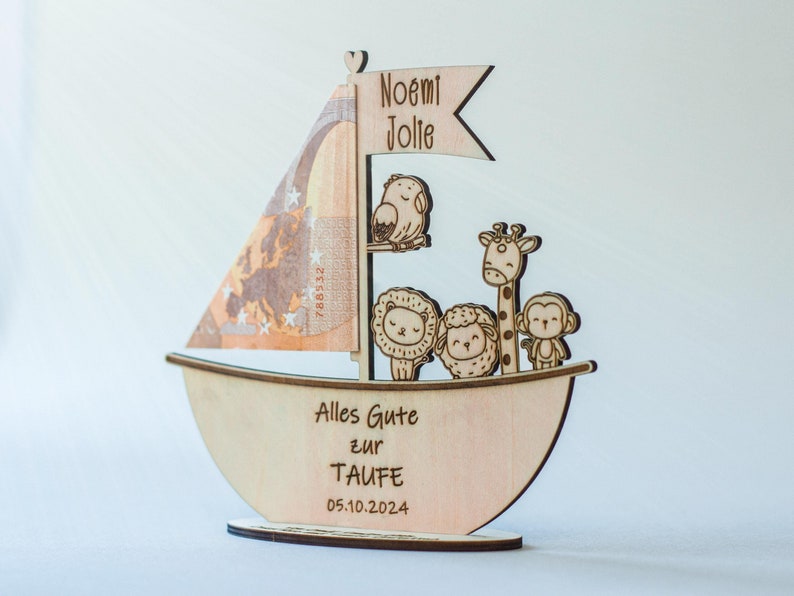 May include: A wooden sailboat with a sail made of a euro bill. The boat is decorated with a giraffe, a lion, a sheep, and a monkey. The boat has the text "Noemi Jolie" on the flag, "Alles Gute zur Taufe" on the side, and "05.10.2024" on the bottom.