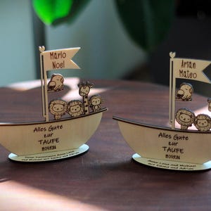 May include: Two wooden sailboats with animal figures, including lions, giraffes, and birds. The sailboats have the text "Alles Gute zur Taufe" and the names "Marlo Noel" and "Artan Mateo".