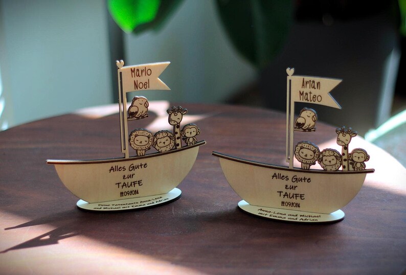 May include: Two wooden sailboats with animal figures, including lions, giraffes, and birds. The sailboats have the text "Alles Gute zur Taufe" and the names "Marlo Noel" and "Artan Mateo".