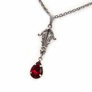 May include: A silver chain necklace with a pendant featuring a detailed ornate design and a dangling red teardrop-shaped gemstone.