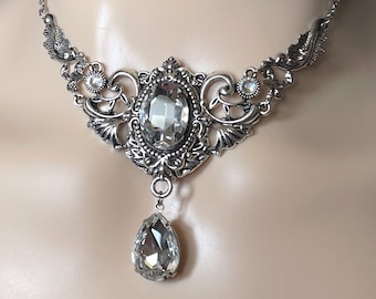 May include: A silver choker necklace with a large, clear crystal pendant. The necklace features intricate, ornate detailing and a smaller crystal drop.