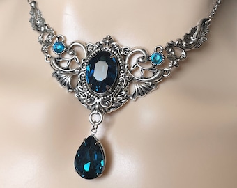 May include: A silver choker necklace with a large blue gemstone pendant and smaller blue gemstones on the sides. The necklace has a delicate, ornate design.