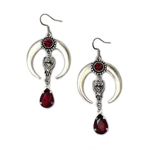 May include: Silver crescent moon earrings with a dangling red teardrop crystal and a small silver bat charm. The earrings have a red rhinestone in the center of the crescent moon.
