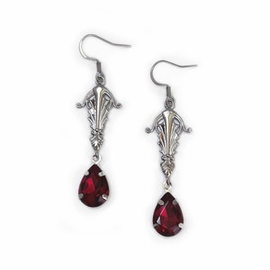 May include: A pair of silver dangle earrings with a teardrop-shaped red gemstone. The earrings have a delicate, ornate design.