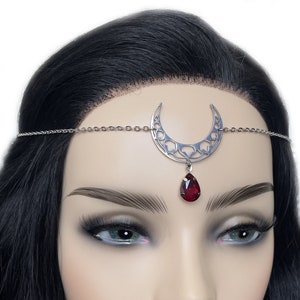 May include: Silver-toned crescent moon headpiece with a dangling teardrop-shaped, deep red gemstone. The moon design has intricate cut-out details. The headpiece is worn on a mannequin with black hair and dark lipstick, against a white background.