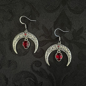 May include: A pair of silver crescent moon earrings with red gemstone accents. The earrings feature intricate Celtic knotwork designs.