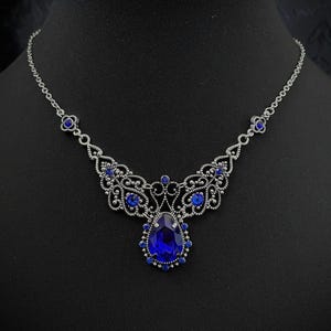 May include: A silver-toned necklace with a large, teardrop-shaped, deep blue gemstone pendant. The necklace features intricate filigree detailing and smaller blue gemstones. The necklace is displayed on a black mannequin.