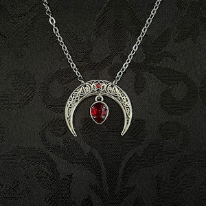 May include: A silver crescent moon necklace with a red gemstone pendant. The pendant is surrounded by intricate details, including a pentagram and other symbols.