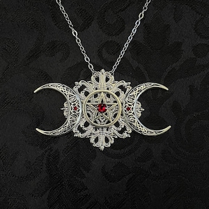 May include: Silver-toned pendant necklace featuring a triple moon design with crescent moons on either side of a central pentagram. The pentagram has a red gemstone in the center. The necklace is on a silver chain.