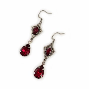 May include: A pair of silver dangle earrings with red teardrop-shaped crystals. The earrings have a delicate design with a small round crystal at the top of each teardrop.