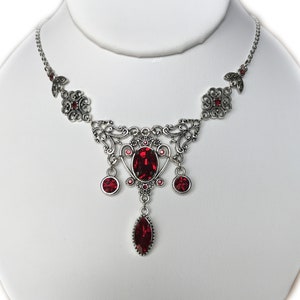 May include: Silver-toned necklace with ornate filigree design, featuring a large oval red gemstone pendant and smaller red gems. The necklace is displayed on a white bust, showcasing its intricate details and vintage style.