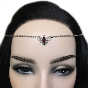 May include: A silver chain forehead crown with a red gemstone in the center. The crown has a delicate, ornate design.