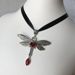 May include: A silver dragonfly pendant necklace with a red crystal. The necklace is on a black ribbon.