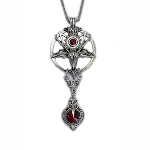 May include: Silver-toned pendant necklace featuring a detailed, gothic-inspired design. The pendant includes a central pentagram with a goat head, adorned with a round, red gemstone. A second, teardrop-shaped red gemstone is at the bottom.