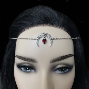May include: Silver crescent moon headpiece with a red gemstone pendant. The moon is decorated with a star and Celtic knotwork.