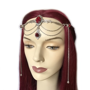 May include: Silver headband with a delicate wing design and red gemstone accents. The headband has dangling chains with small red gemstones at the ends.