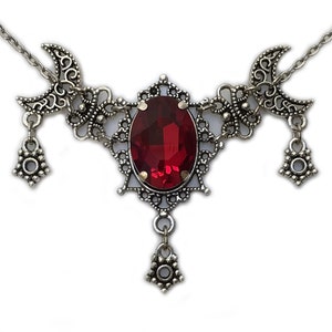 May include: A silver necklace with a large red gemstone pendant and two crescent moon charms. The pendant is surrounded by intricate filigree details and the necklace has dangling charms.