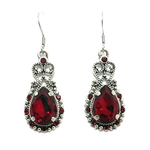 May include: A pair of silver-toned drop earrings featuring a large, central pear-shaped red gemstone surrounded by a border of smaller red crystals. The metalwork includes intricate filigree detailing above the main stone and a hook-style closure.