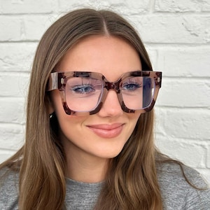 May include: Square-framed spectacles with a tortoiseshell pattern. The glasses have a thick frame and clear lenses. The image is a close-up of the spectacles, with a person wearing them. The background is a white brick wall.