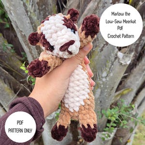 May include: A handmade crochet meerkat toy in shades of brown, tan, and cream. The plush animal has a textured, fuzzy appearance and is held in a person's hand. Text on the image reads "Marlow the Low-Sew Meerkat PDF Crochet Pattern" and "PDF Pattern Only."