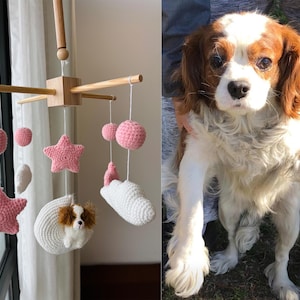 Op de afbeelding: Een houten babymobiel met gehaakte roze sterren, roze bollen, witte wolken en een witte halve maan met een kleine bruin-witte hondfiguur eraan bevestigd. De mobiel hangt aan een centraal houten kruisframe met witte koordjes.