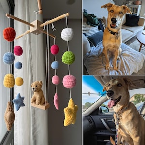 Op de afbeelding: Een mobiel met een centraal houten kruis en hangende witte koorden. Hieraan zijn diverse gehaakte vormen bevestigd, waaronder bollen in rood, blauw, geel, wit, groen en roze, evenals blauwe, bruine en gele sterren en een centrale teddybeer.