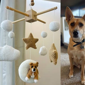 Op de afbeelding: Een hangende mobiel met een houten kruisframe, witte gehaakte wolken, beige gehaakte sterren en een witte gehaakte maan met een kleine bruin-witte hondenfiguur. Daarnaast zit een beige hond met puntige oren en een zwarte halsband.