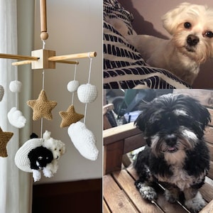 Op de afbeelding: Een baby-mobiel met een houten kruisframe en hangende gehaakte elementen, waaronder witte bollen, witte wolken, beige sterren en twee kleine gehaakte hondjes, &eacute;&eacute;n wit en &eacute;&eacute;n zwart-wit, opgehangen aan witte koordjes.