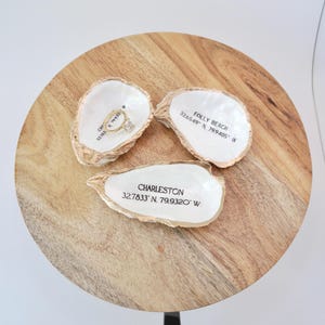 May include: Three white oyster shell ring dishes with gold accents. The dishes are labeled with the names of locations and their coordinates: Charleston 32.7833' N 79.9320' W, Folly Beach 32.6549' N 79.9405' W, and CHA 32783 19933. A gold ring is sitting in the Charleston dish.