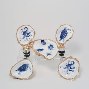 May include: Set of five oyster shell bottle stoppers with blue and white painted designs. The designs include an octopus, a turtle, and a seashell with a starfish. The stoppers are gold-trimmed.