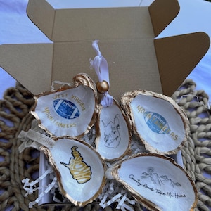May include: Set of four oyster shell ornaments with gold accents. The ornaments feature West Virginia-themed designs, including the state outline, a football, and the words "West Virginia Mountaineers". One ornament features the text "Country Roads Take Me Home".