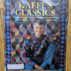 May include: A book cover featuring the title "KAFFE'S CLASSICS" in yellow letters. The cover shows a smiling man wearing a colorful sweater. The book is about knitting patterns for sweaters, jackets, and vests. The author's name, Kaffe Fassett, is also displayed.