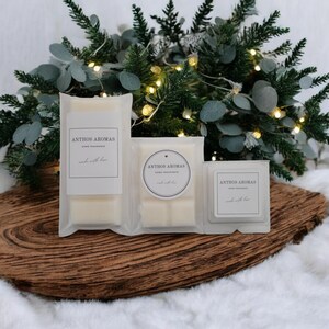 May include: Three white wax melts in clear plastic packaging. The wax melts are labelled "Anthos Aromas" and "Made with Love". The packaging is surrounded by a green garland with white lights.