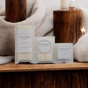 May include: Three Anthos Aromas wax melt packages on a wooden surface. The clear packages display white wax melts. Each package features a label with the brand name and the phrase "made with love."