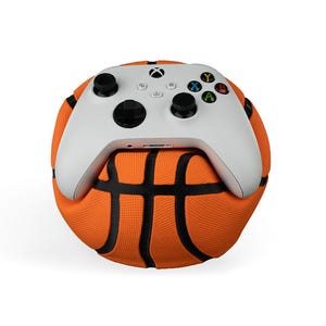 Puede incluir: Un mando blanco de Xbox descansa sobre una pelota de baloncesto naranja. El mando tiene joysticks negros y botones de colores. La pelota de baloncesto tiene líneas negras y una superficie texturizada.