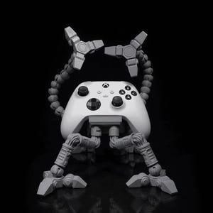 May include: A white Xbox controller with a gray, articulated stand. The stand has four legs and two arms that extend from the top of the controller. The arms are positioned in a way that suggests the controller is holding a weapon.