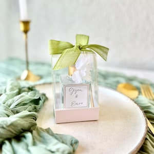 Può includere: Un flacone diffusore in vetro trasparente con tappo dorato e fiori bianchi, presentato in una scatola trasparente con un nastro verde chiaro. L'etichetta recita "Gizem & Emre". La scatola è su un piatto bianco con un panno verde.