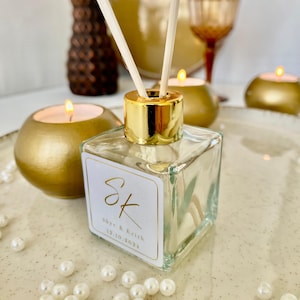 Può includere: Una bottiglia di diffusore in vetro trasparente con tappo dorato e bastoncini. La bottiglia ha un'etichetta bianca con il testo "SK Skye & Keith 12.10.2024". La bottiglia è circondata da perle bianche.