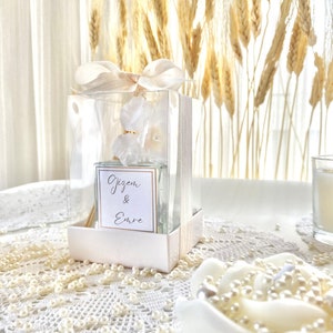Op de afbeelding: Een witte geschenkdoos met een doorzichtig deksel, met daarin een witte diffuser met een bloemenmotief. De doos is vastgebonden met een wit lint en heeft de namen "Gizem & Emre" erop staan. De doos staat op een witte tafel met een wit kantkleed en witte parels.