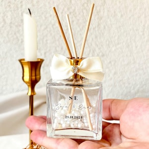 May include: A clear glass diffuser bottle filled with white pearls and wooden reeds. The bottle is adorned with a cream-colored bow and a decorative jewel. The bottle has the initials "SE" and the names "Seral & Enes" and the date "29.11.2021" printed on it.