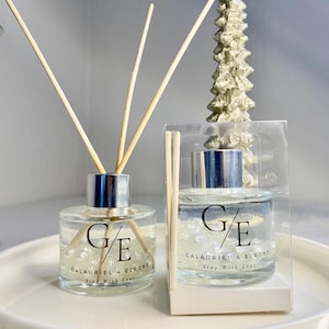 May include: Two clear glass bottles with silver caps and black labels. The labels read "G/E GALADRIEL & ELROND Stay With Love". The bottles are filled with a clear liquid and have wooden sticks inserted into them. The bottles are sitting on a white surface.