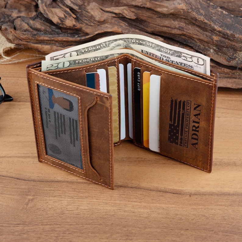 May include: A brown leather bifold wallet featuring a clear ID window, multiple card slots, and a cash compartment containing US currency. The exterior is engraved with an American flag graphic and the text PRIVATE PROPERTY OF ADRIAN.