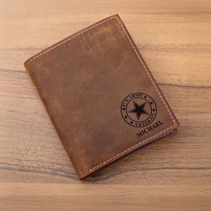 May include: A brown leather passport holder featuring a distressed finish and light brown stitching. The front is engraved with a circular U.S. ARMY VETERAN emblem containing a central star, with the name MICHAEL printed in black text below it.