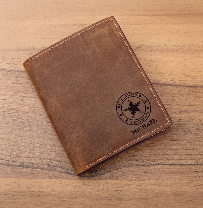 May include: A brown leather passport holder featuring a distressed finish and light brown stitching. The front is engraved with a circular U.S. ARMY VETERAN emblem containing a central star, with the name MICHAEL printed in black text below it.