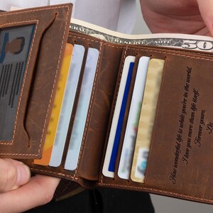 May include: A brown leather bi-fold wallet featuring a clear ID window slot, multiple card slots, and a bill compartment containing a fifty-dollar bill. The interior right panel is engraved with the text: How wonderful life is while you're in the world. I love you more than life itself. Forever yours, -Liz.