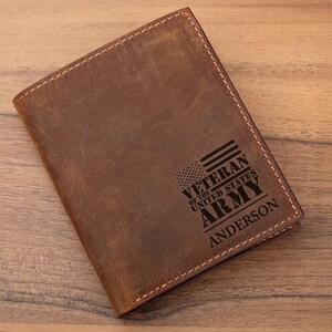 May include: A brown leather passport holder featuring a laser-engraved design of an American flag and the text VETERAN OF THE UNITED STATES ARMY ANDERSON. The cover has visible stitching along the edges and a distressed, matte finish texture.