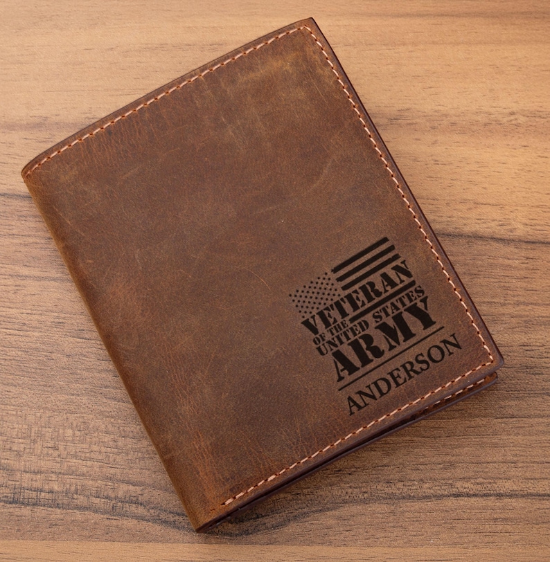 May include: A brown leather passport holder featuring a laser-engraved design of an American flag and the text VETERAN OF THE UNITED STATES ARMY ANDERSON. The cover has visible stitching along the edges and a distressed, matte finish texture.
