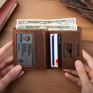 May include: A brown leather bifold wallet featuring a clear ID window on the left and multiple card slots on the right. The wallet contains US currency, including a fifty and a twenty dollar bill, and is engraved with the text Land of the Free Because of the Brave and the name Wilson.