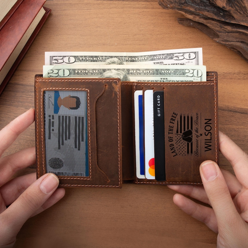 May include: A brown leather bifold wallet featuring a clear ID window on the left and multiple card slots on the right. The wallet contains US currency, including a fifty and a twenty dollar bill, and is engraved with the text Land of the Free Because of the Brave and the name Wilson.