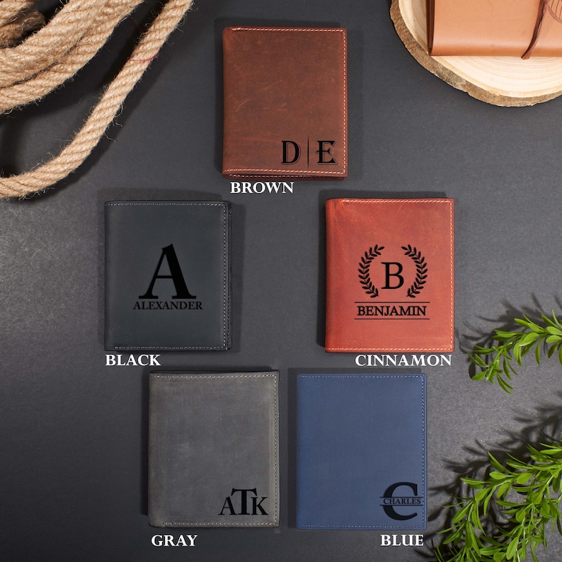 May include: Five rectangular leather wallets in brown, black, cinnamon, gray, and blue. Each wallet features a unique black laser-engraved monogram or name, including the letters D E, A Alexander, B Benjamin, AtK, and C Charles, with visible stitching.